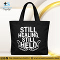 "Still Healing. Still Held." Christian Tote Bag – Durable 12oz Cotton Canvas Reusable Shoulder Bag for Daily Use